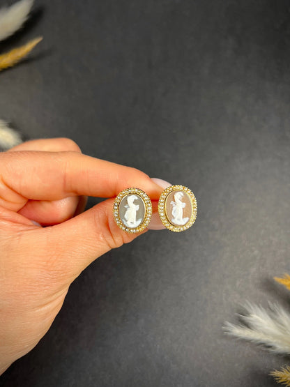 A pair of yellow gold oval shell cameo earrings, held in a hand