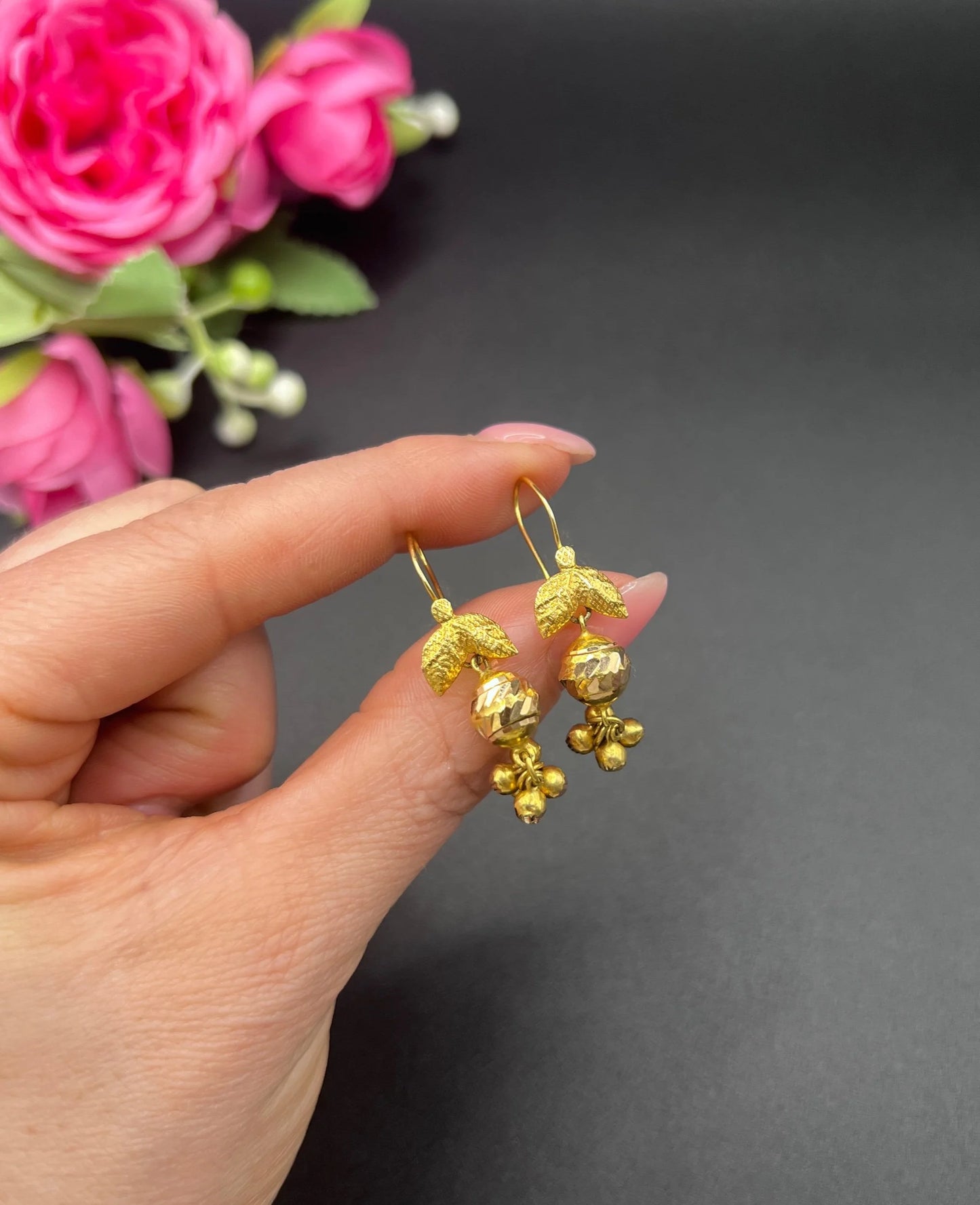 A pair of yellow gold leaf and faceted ball drop earrings with hook fittings, held in a hand