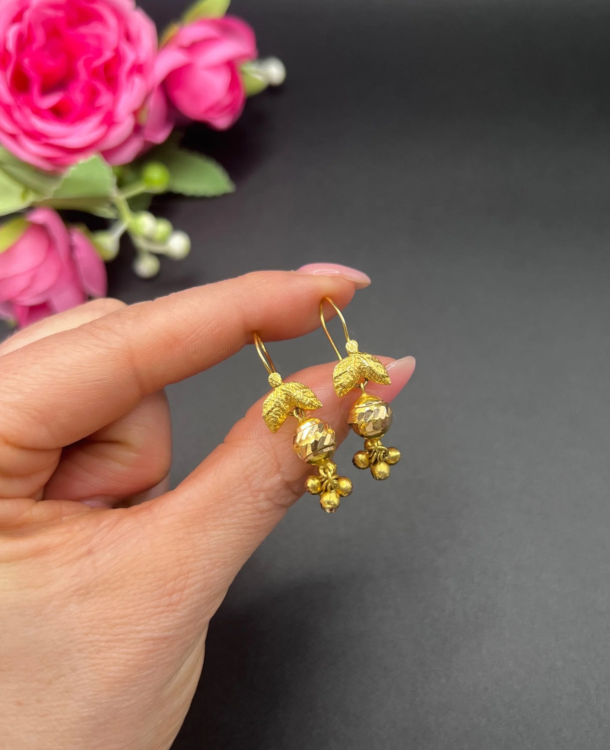 A pair of yellow gold leaf and faceted ball drop earrings with hook fittings, held in a hand