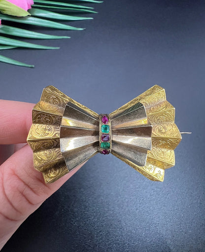 A yellow gold bow shaped brooch with rubies and emeralds, held in a hand