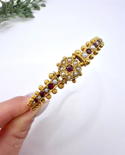 A yellow gold bangle bracelet set with rubies and seed pearls, held in a hand