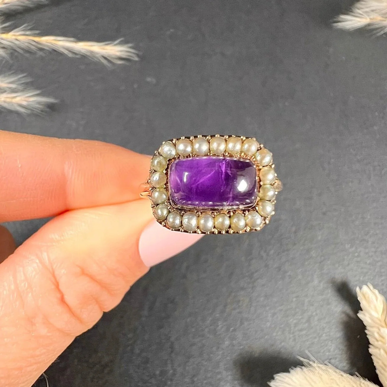 A cabochon amethyst and pearl halo cluster ring in gold, held in a hand