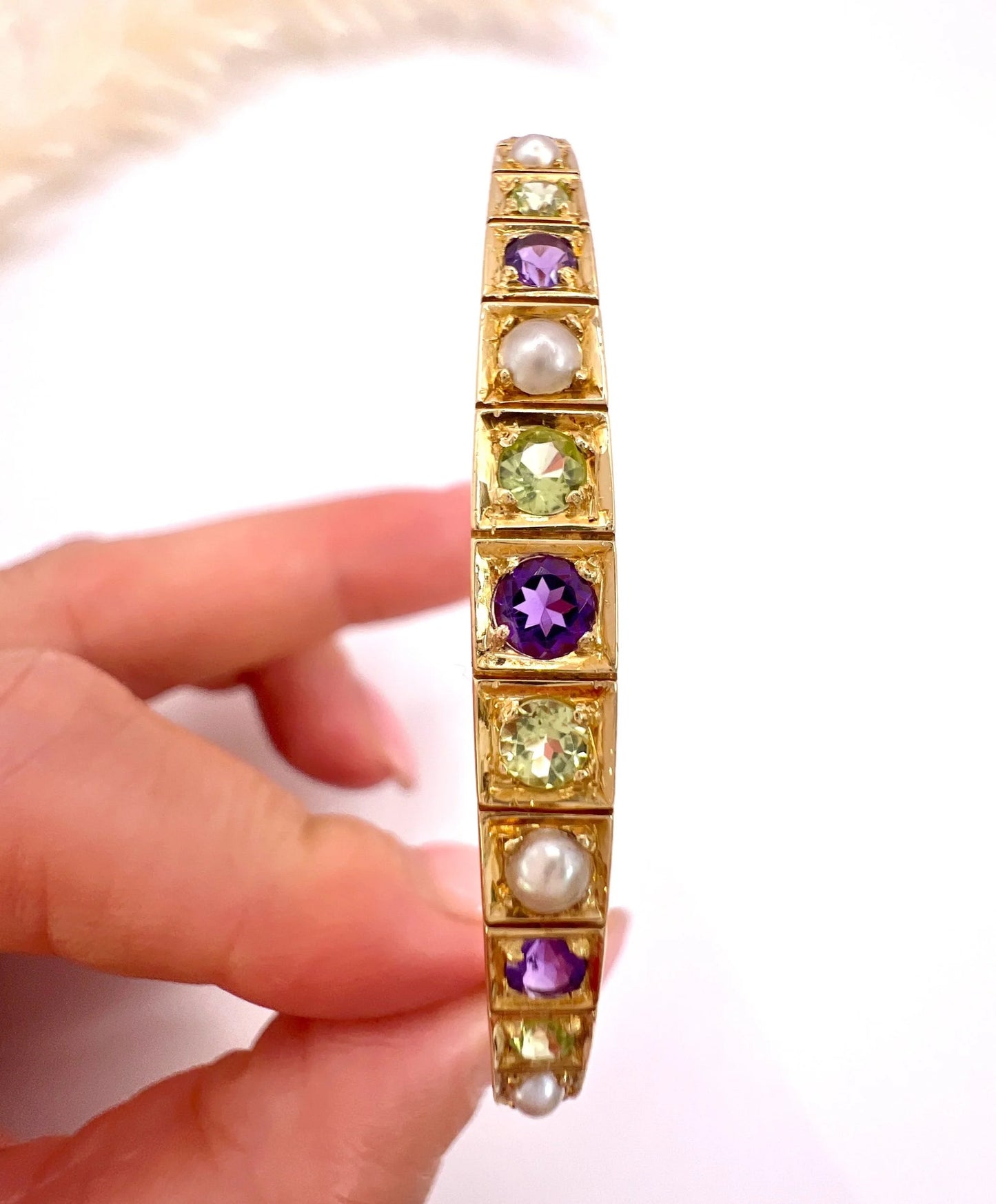 A yellow gold peridot, amethyst and pearl bangle bracelet, held in a hand