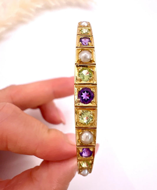 A yellow gold peridot, amethyst and pearl bangle bracelet, held in a hand