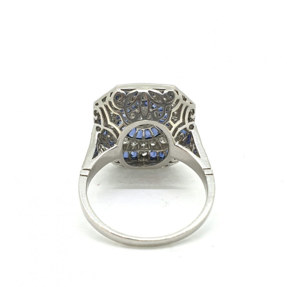Diamond and Calibre Sapphire Target Cluster Dress Ring in Platinum; central diamond within a calibre sapphire halo, surrounded by brilliant-cut diamonds and a further outer border of sapphires, with diamond and sapphire set shoulders and a beautifully pierced under-gallery