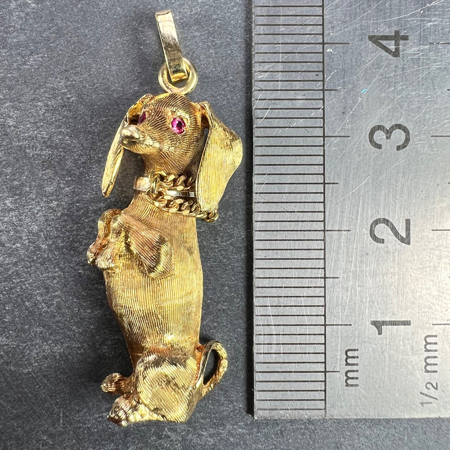 A yellow gold dachshund dog charm with ruby eyes, next to a metal ruler on a grey background