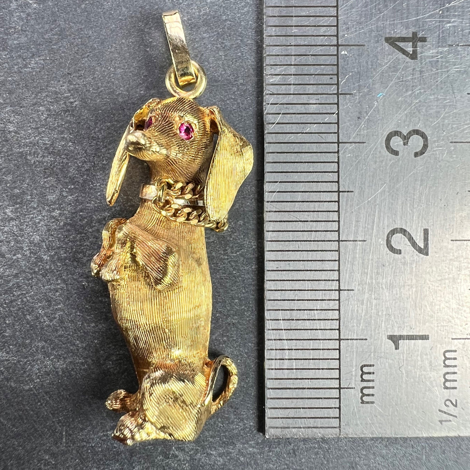 A yellow gold dachshund dog charm with ruby eyes, next to a metal ruler on a grey background