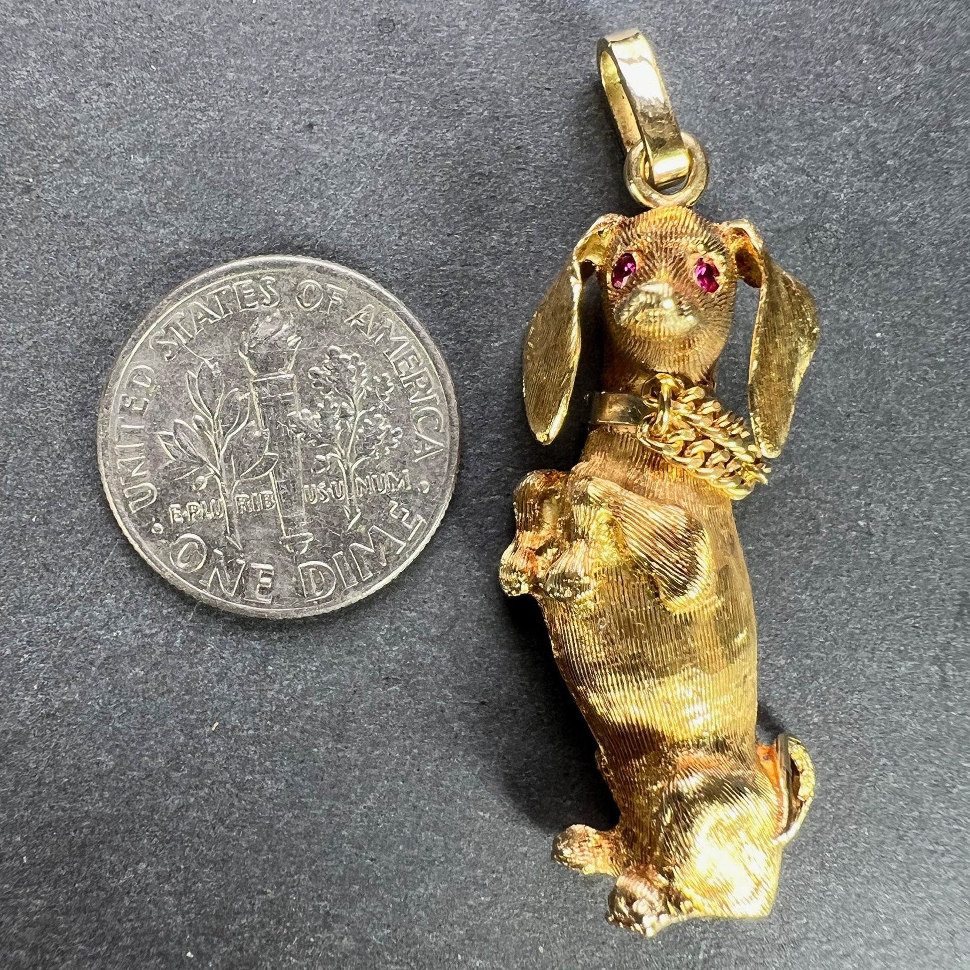 A yellow gold dachshund dog charm with ruby eyes, next to a dime coin on a grey background