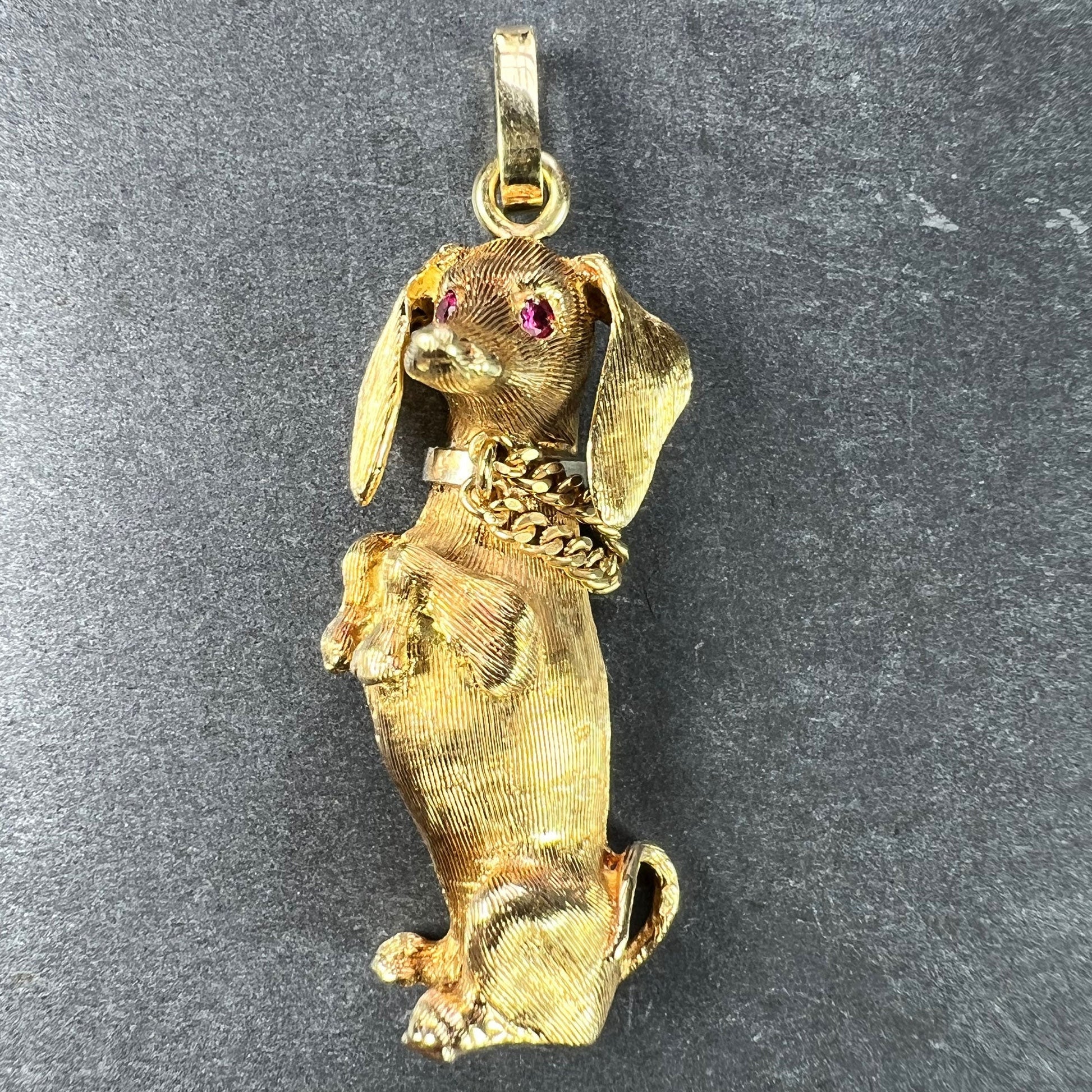 A yellow gold dachshund dog charm with ruby eyes, on a dark background
