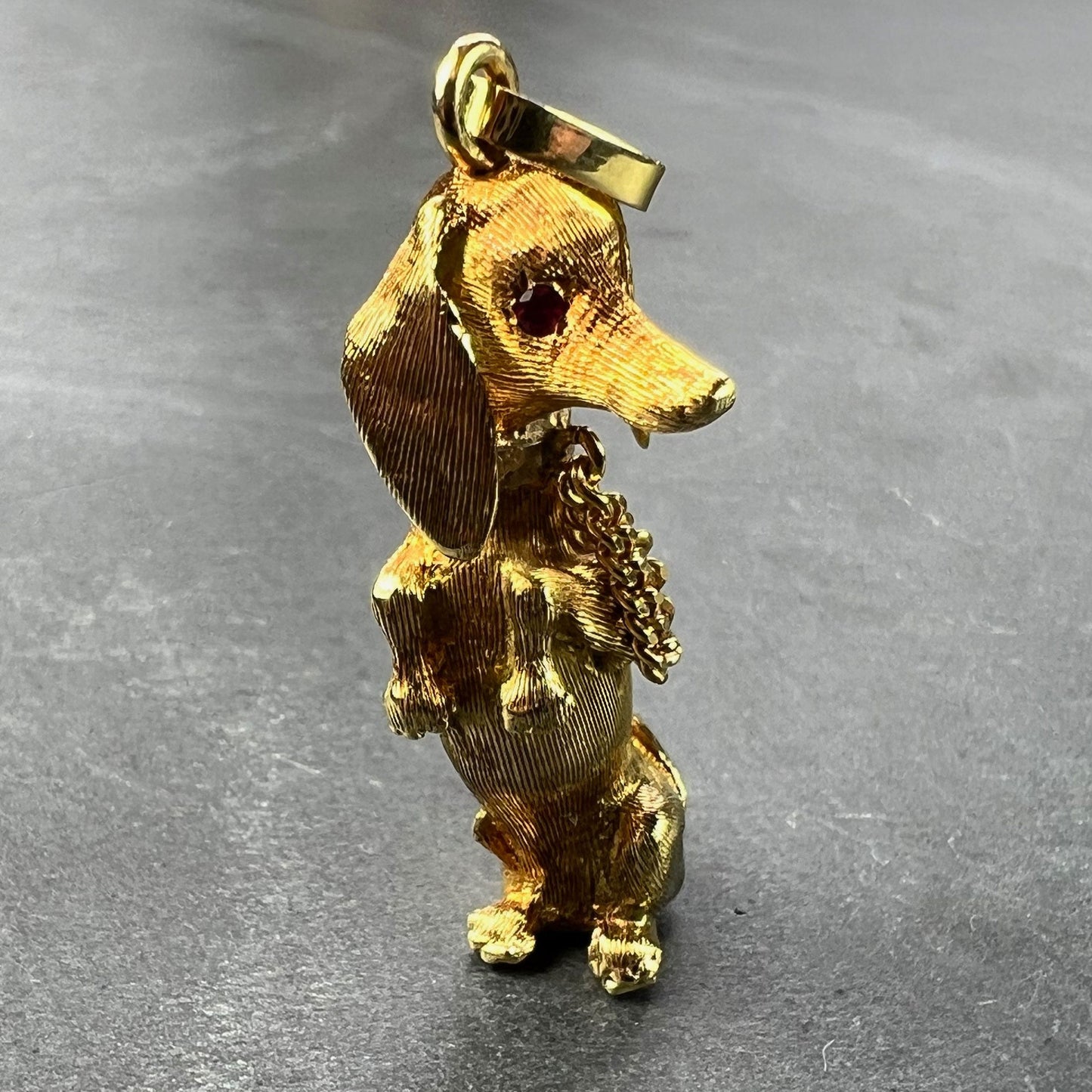 A yellow gold dachshund dog charm with ruby eyes, on a grey background