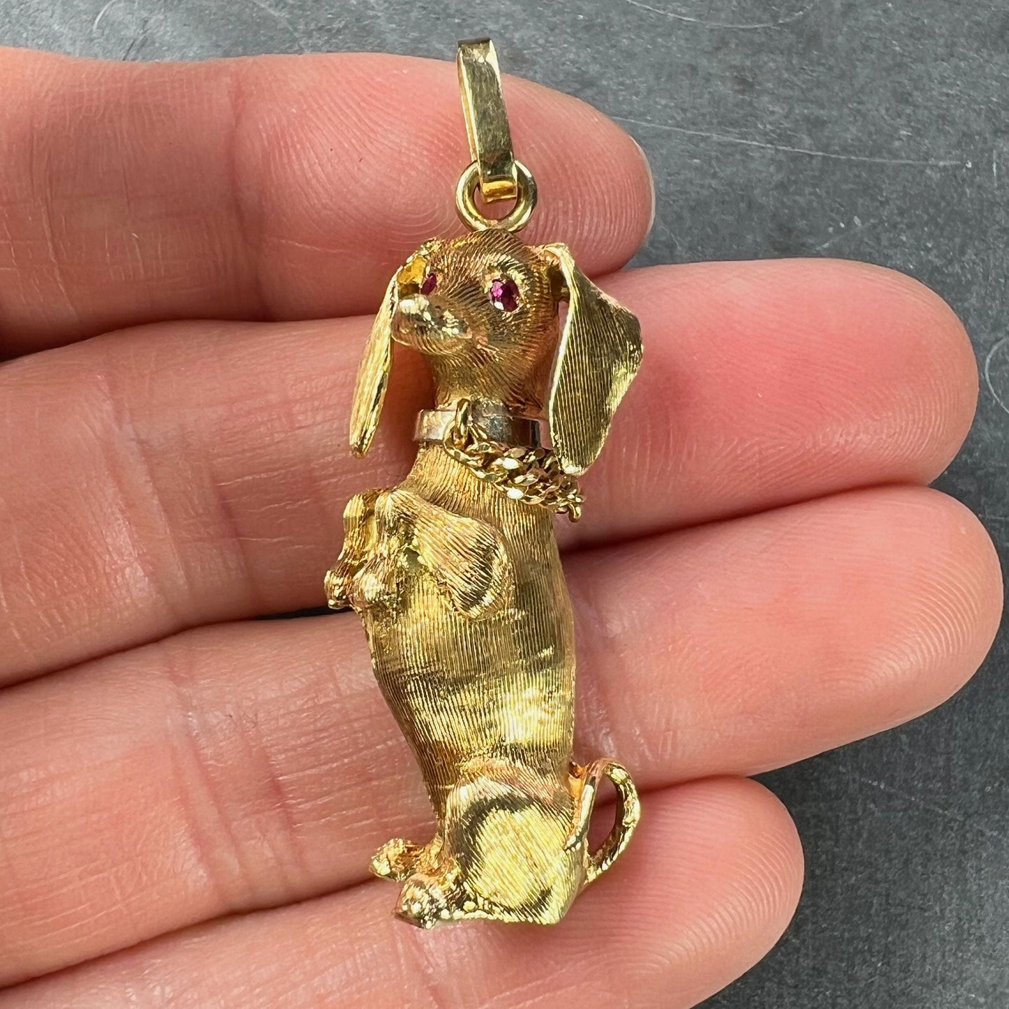 A yellow gold dachshund dog charm with ruby eyes, held in a hand on a grey background