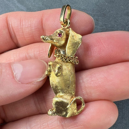 A yellow gold dachshund dog charm with ruby eyes, held in a hand on a grey background