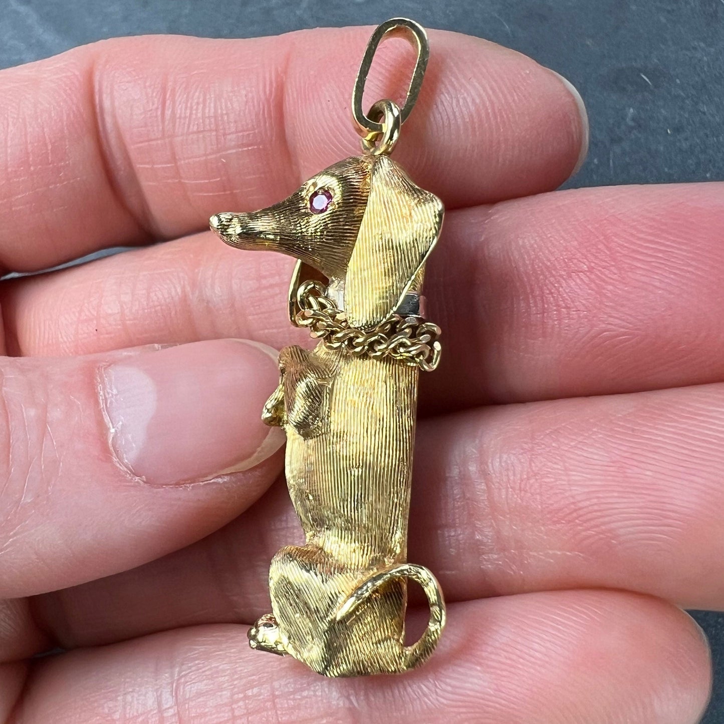 A yellow gold dachshund dog charm with ruby eyes, held in a hand on a grey background