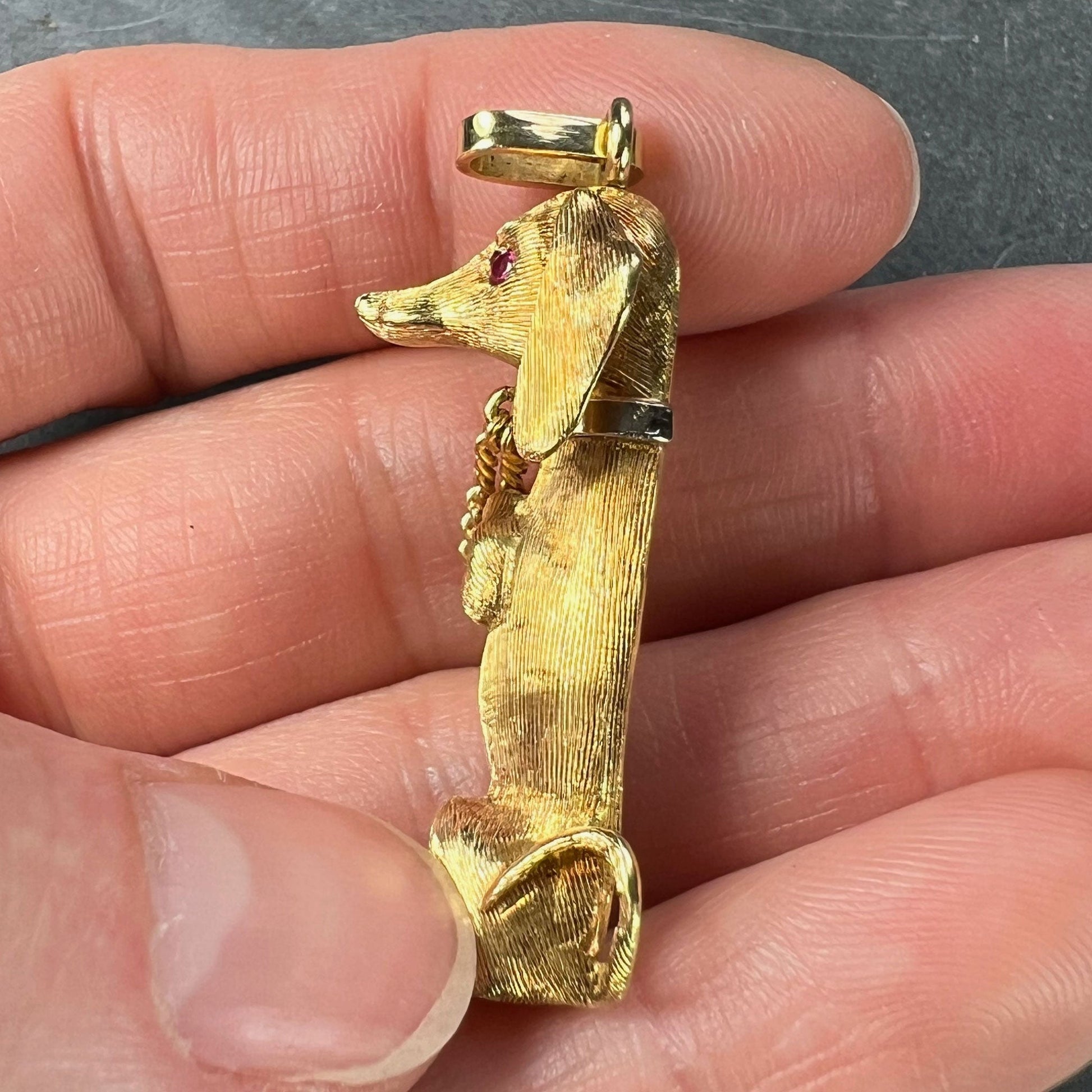 A yellow gold dachshund dog charm with ruby eyes, held in a hand on a grey background