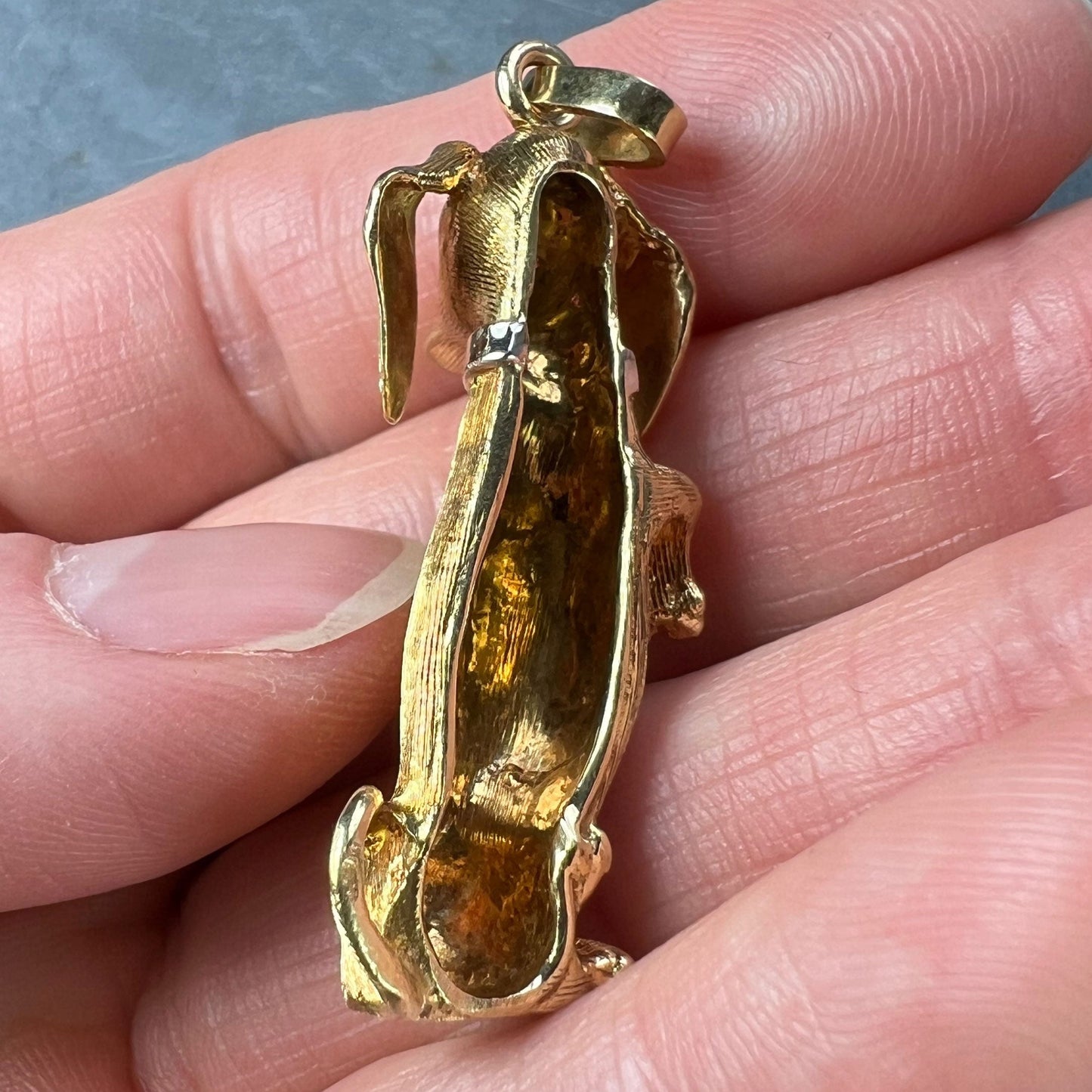 A yellow gold dachshund dog charm with ruby eyes, held in a hand on a grey background