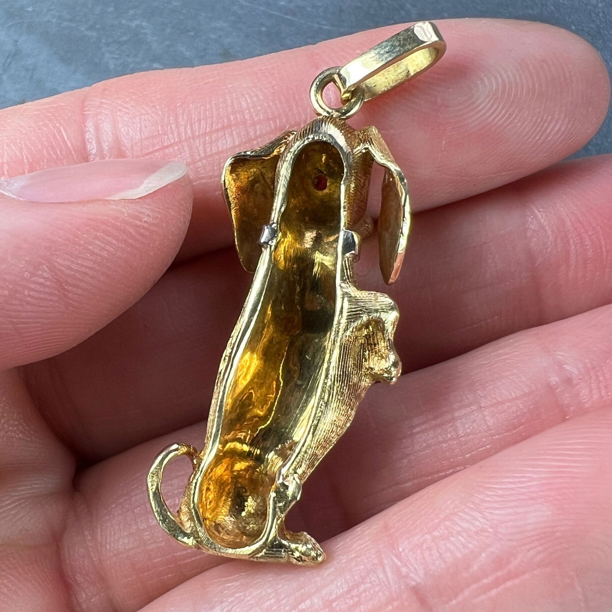 A yellow gold dachshund dog charm with ruby eyes, held in a hand on a grey background