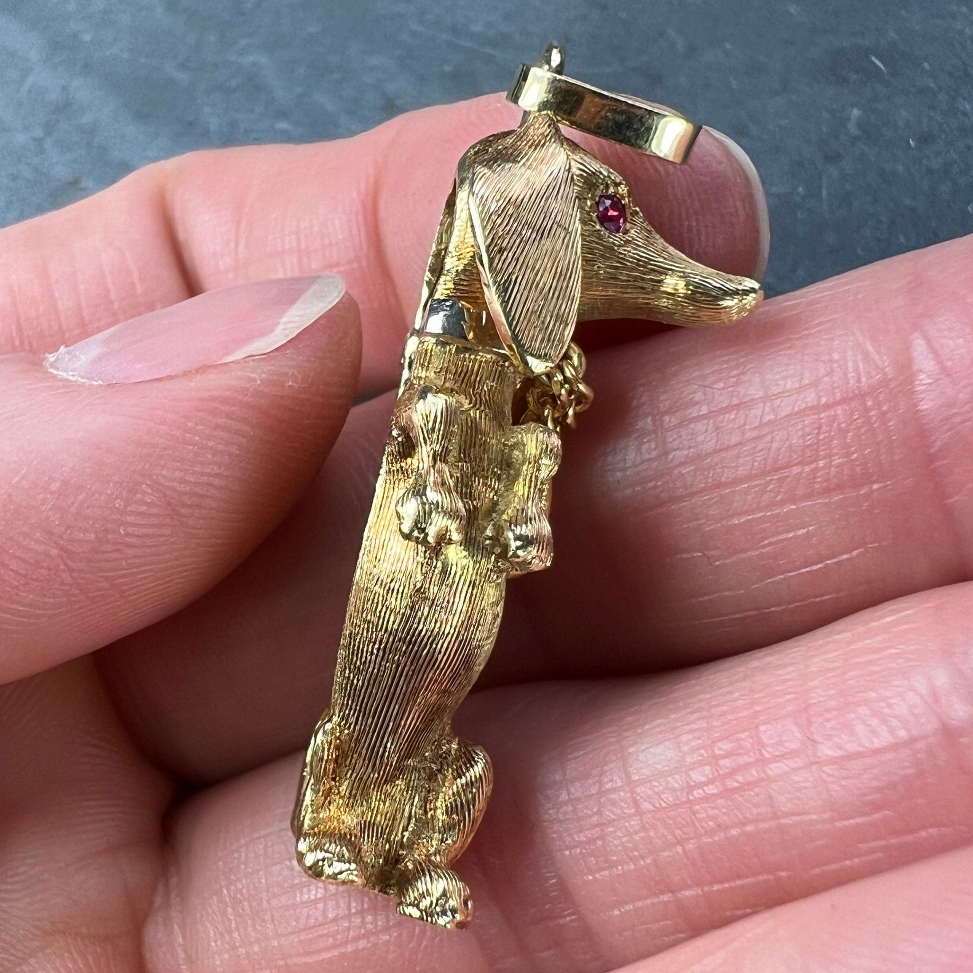 A yellow gold dachshund dog charm with ruby eyes, held in a hand on a grey background