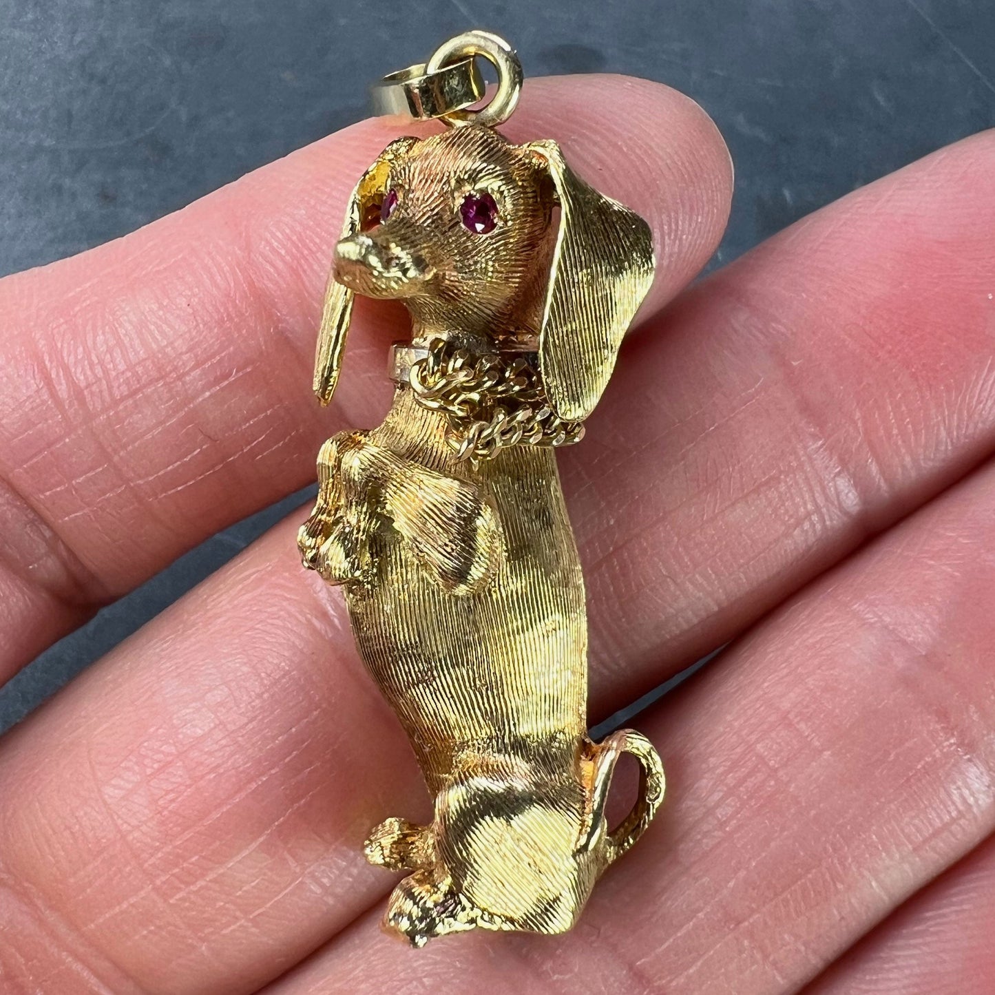 A yellow gold dachshund dog charm with ruby eyes, held in a hand on a grey background