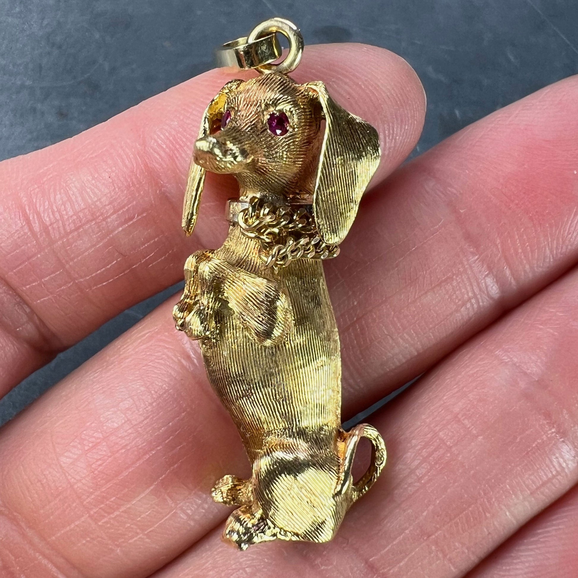 A yellow gold dachshund dog charm with ruby eyes, held in a hand on a grey background