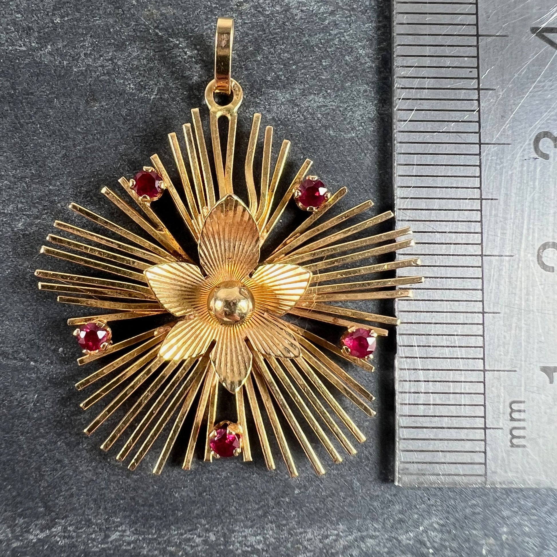 A yellow gold floral sunburst pendant with rubies, next to a metal ruler on a grey background