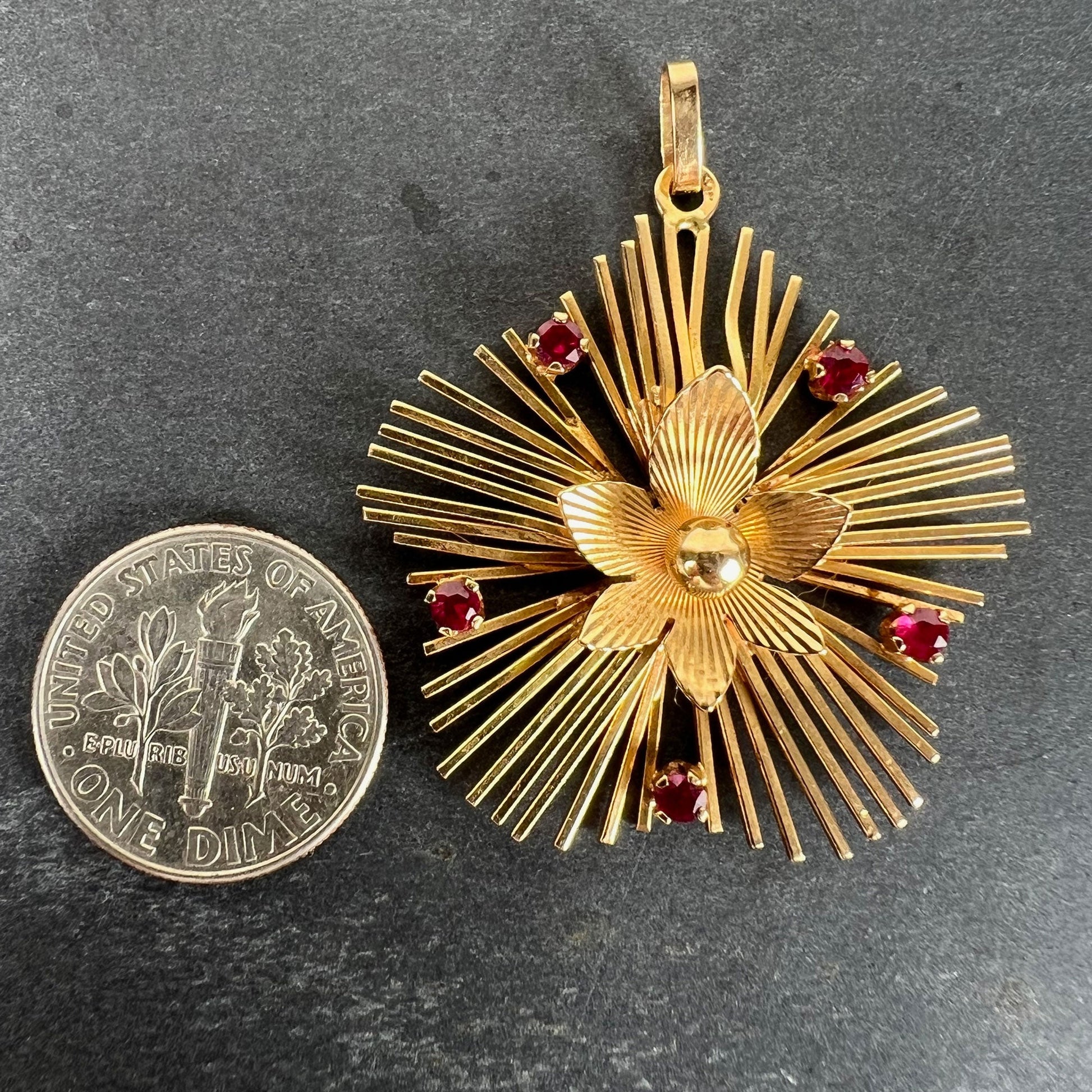 A yellow gold floral sunburst pendant with rubies, next to a dime coin on a grey background