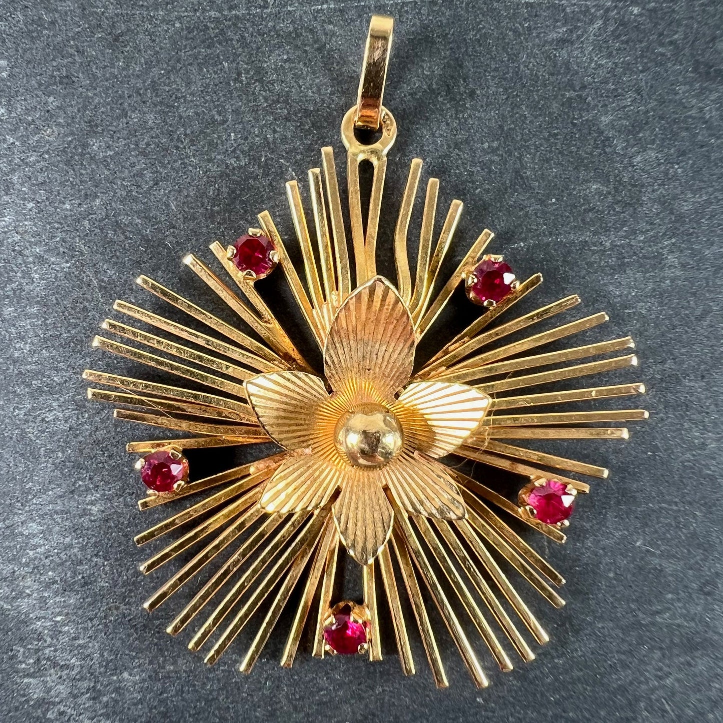 A yellow gold floral sunburst pendant with rubies, on a grey background