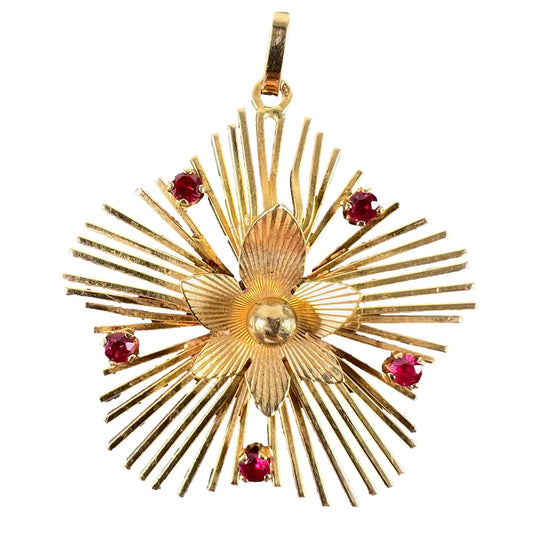 A yellow gold floral sunburst pendant with rubies, on a white background