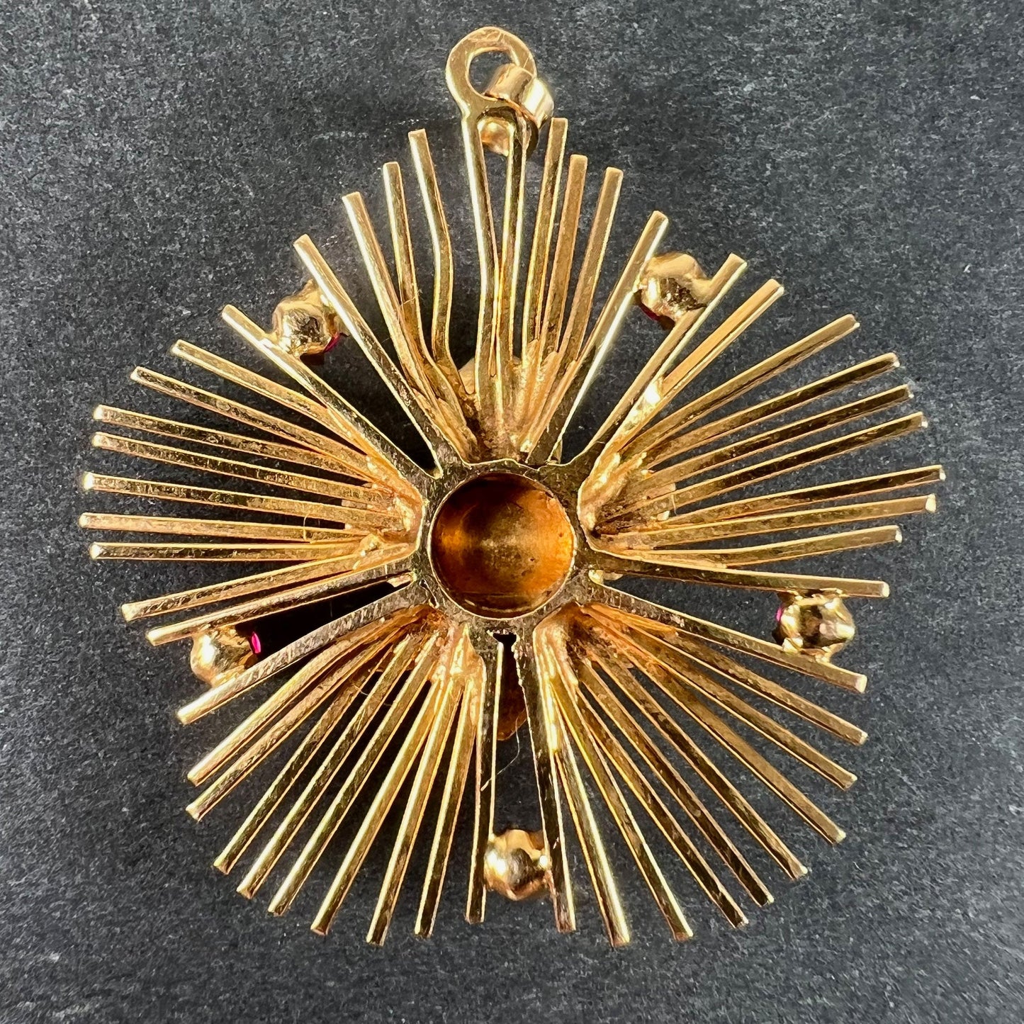 A yellow gold floral sunburst pendant with rubies, on a grey background