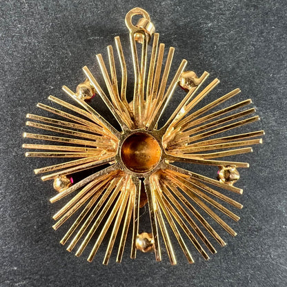 A yellow gold floral sunburst pendant with rubies, on a grey background