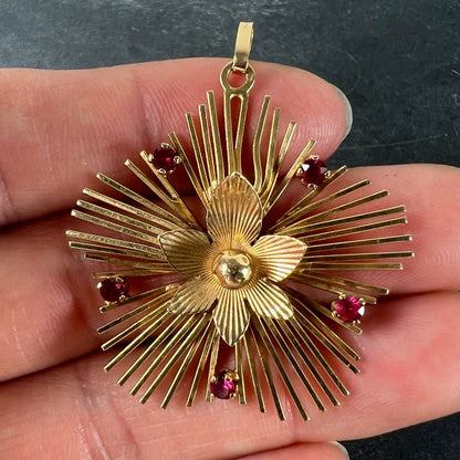 A yellow gold floral sunburst pendant with rubies, held in a hand on a grey background