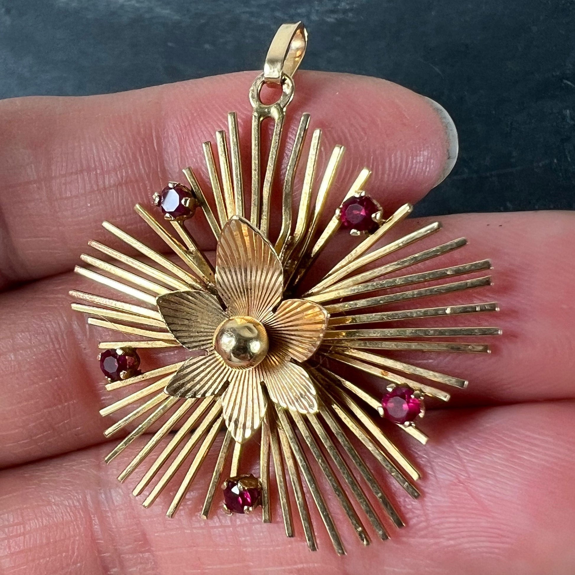 A yellow gold floral sunburst pendant with rubies, held in a hand on a grey background