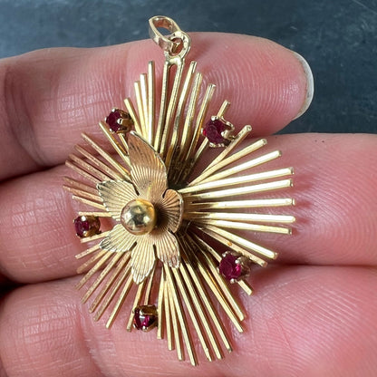 A yellow gold floral sunburst pendant with rubies, held in a hand on a grey background