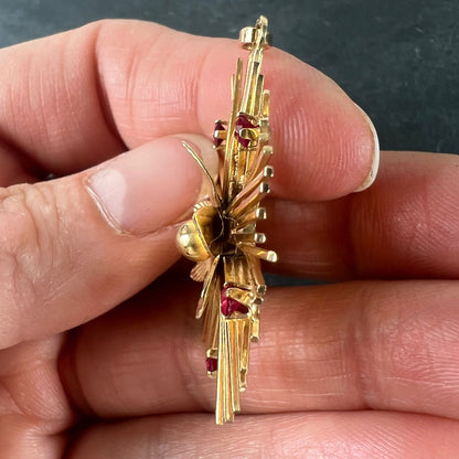 A yellow gold floral sunburst pendant with rubies, held in a hand on a grey background