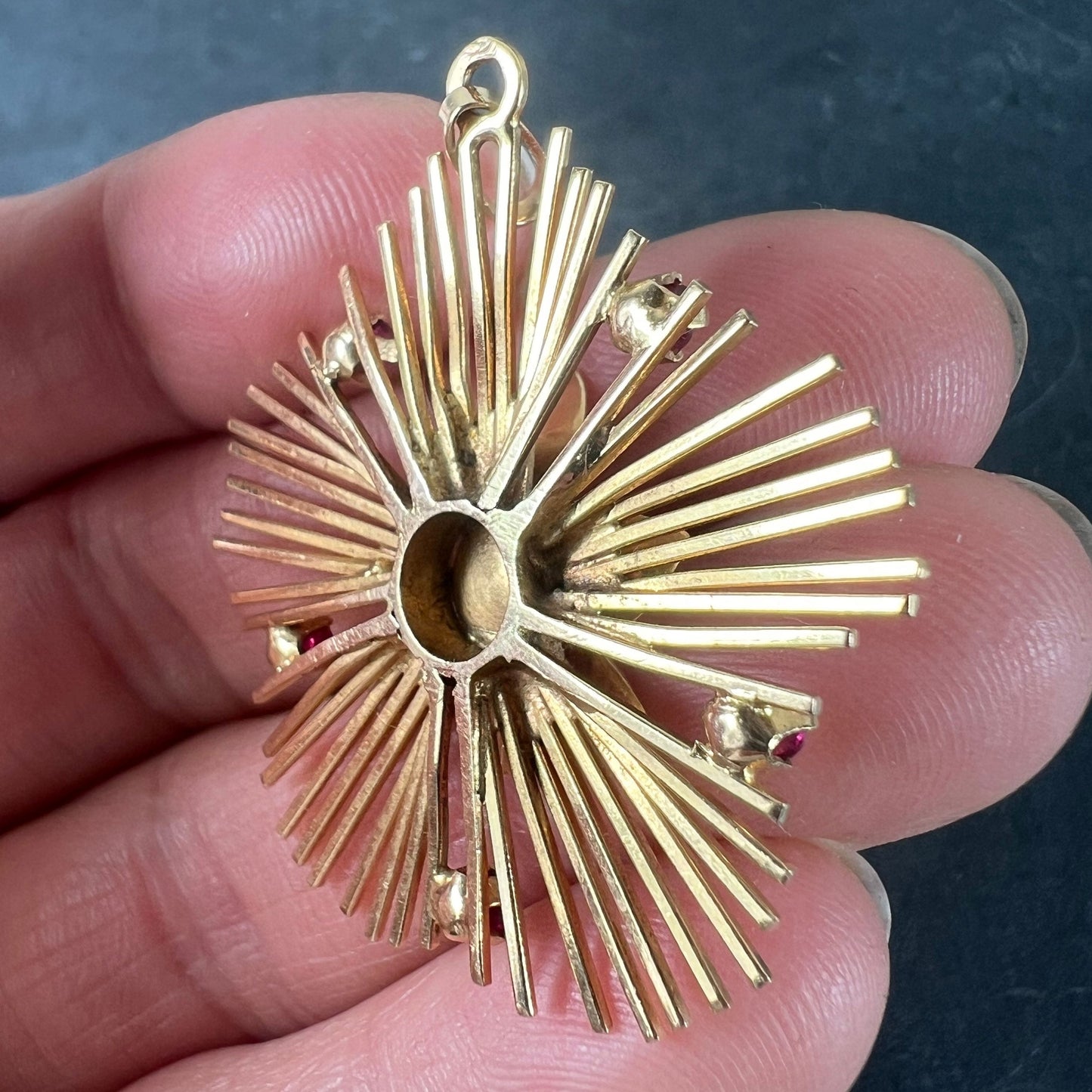 A yellow gold floral sunburst pendant with rubies, held in a hand on a grey background