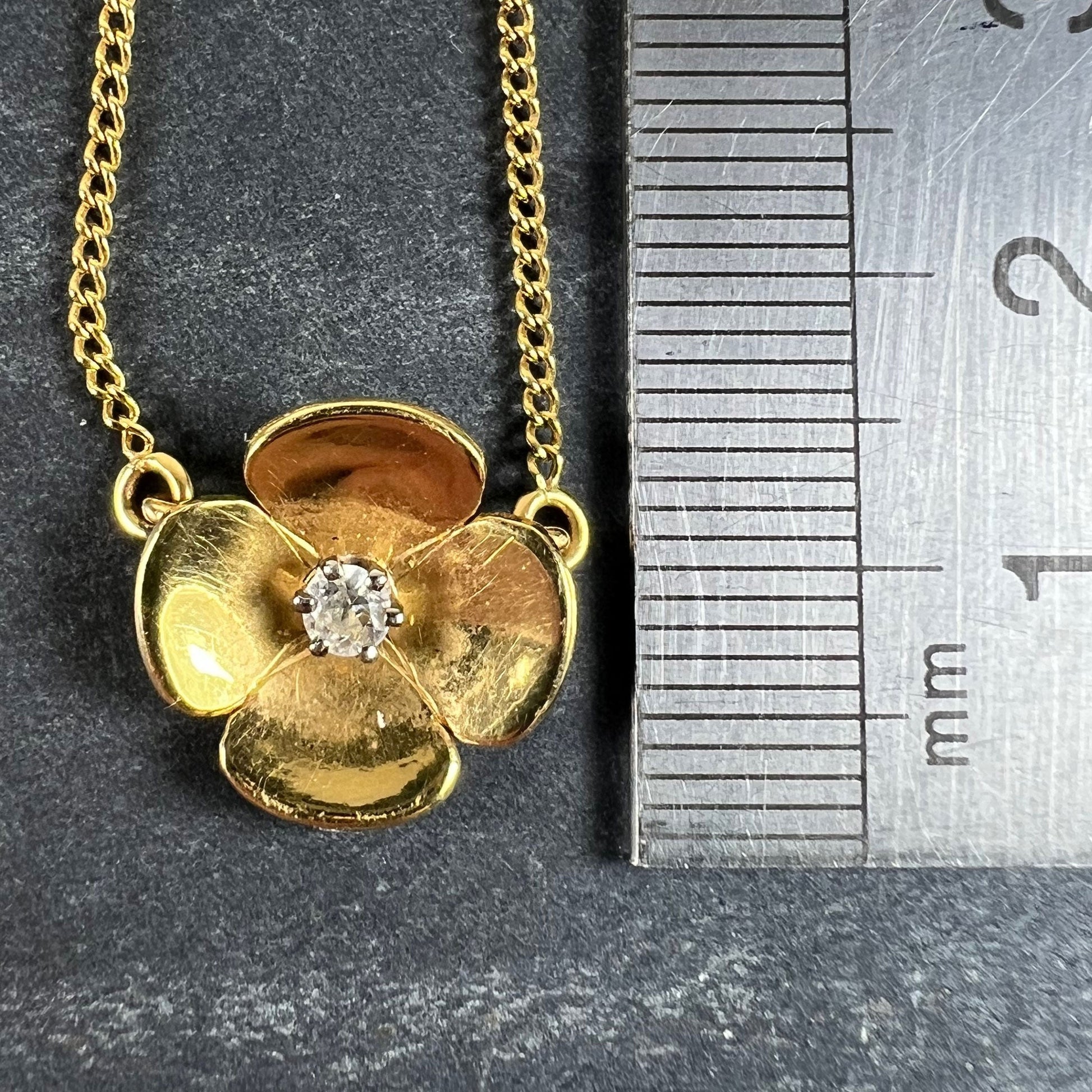 A yellow gold and diamond flower pendant necklace, next to a metal ruler on a grey background