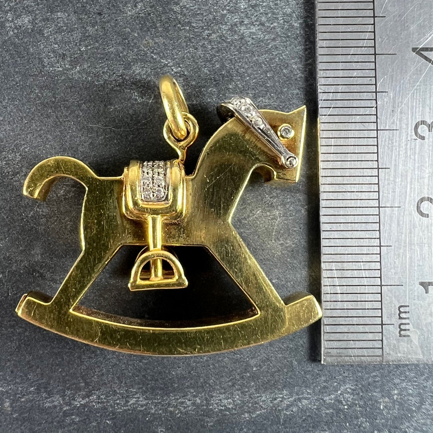 A yellow and white gold rockinghorse pendant with diamonds, next to a metal ruler on a grey background