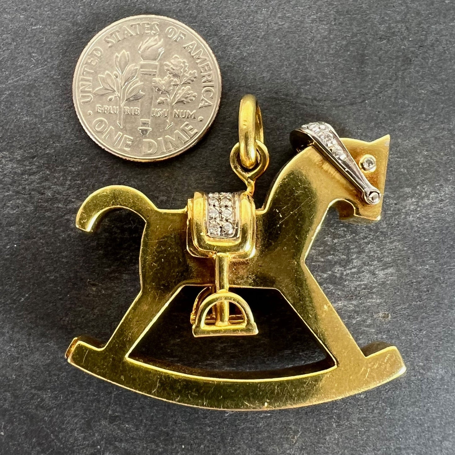 A yellow and white gold rockinghorse pendant with diamonds, next to a dime coin on a grey background