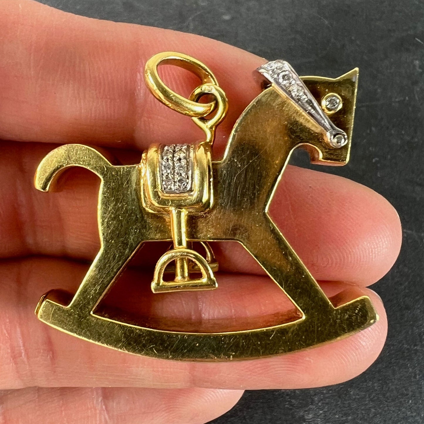 A yellow and white gold rockinghorse pendant with diamonds, held in a hand on a grey background