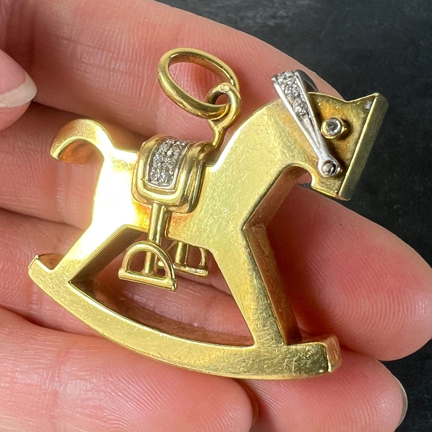 A yellow and white gold rockinghorse pendant with diamonds, held in a hand on a grey background