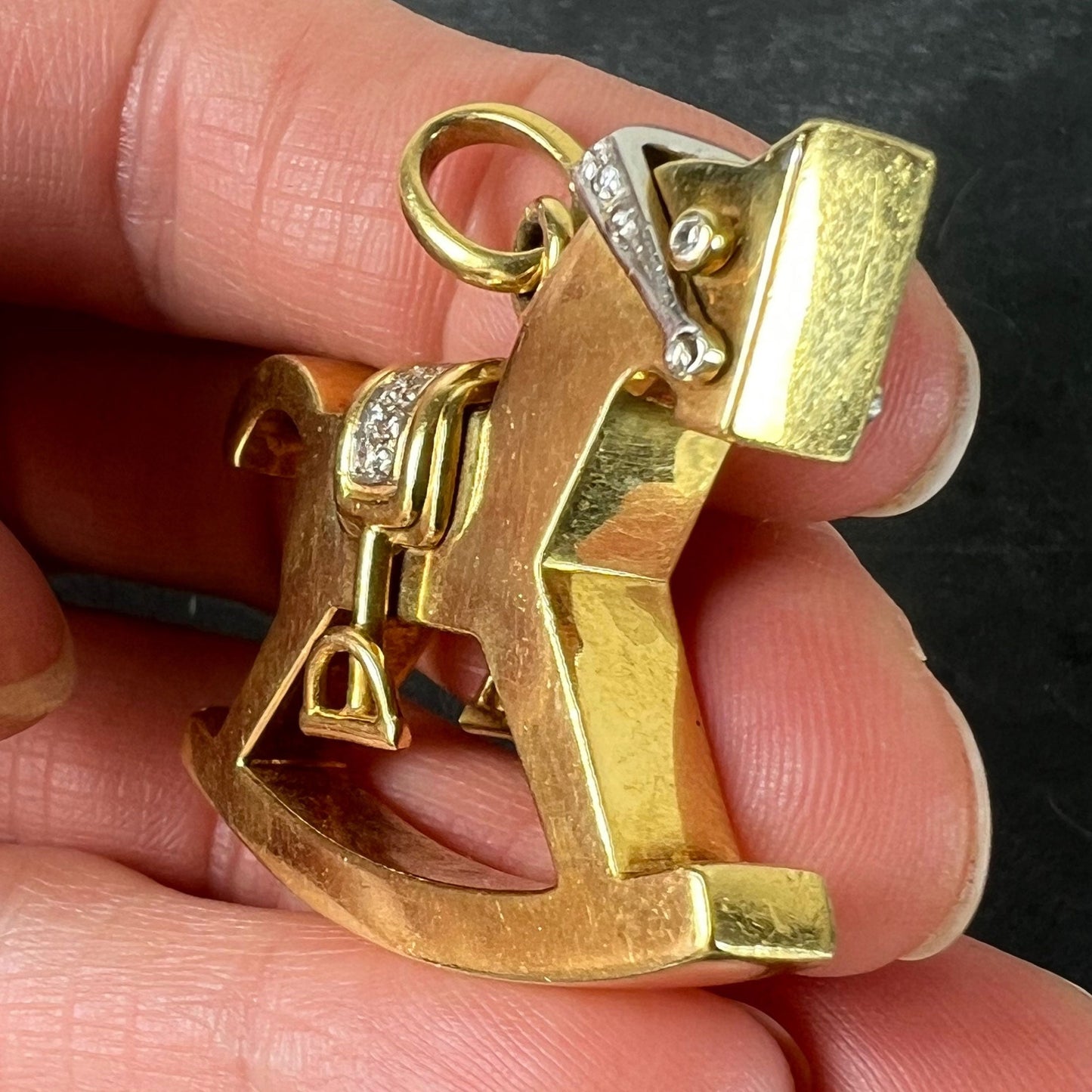 A yellow and white gold rockinghorse pendant with diamonds, held in a hand on a grey background