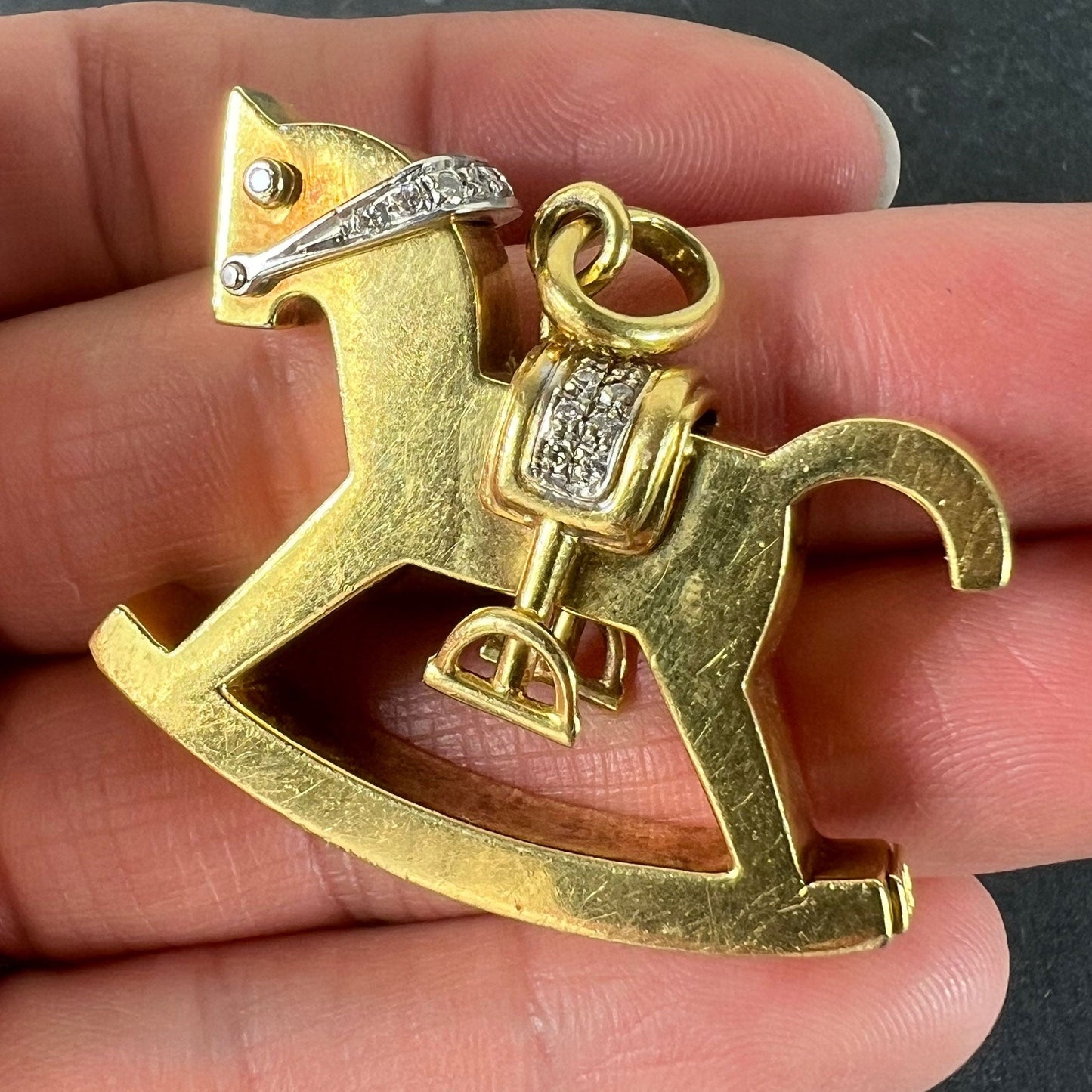 A yellow and white gold rockinghorse pendant with diamonds, held in a hand on a grey background