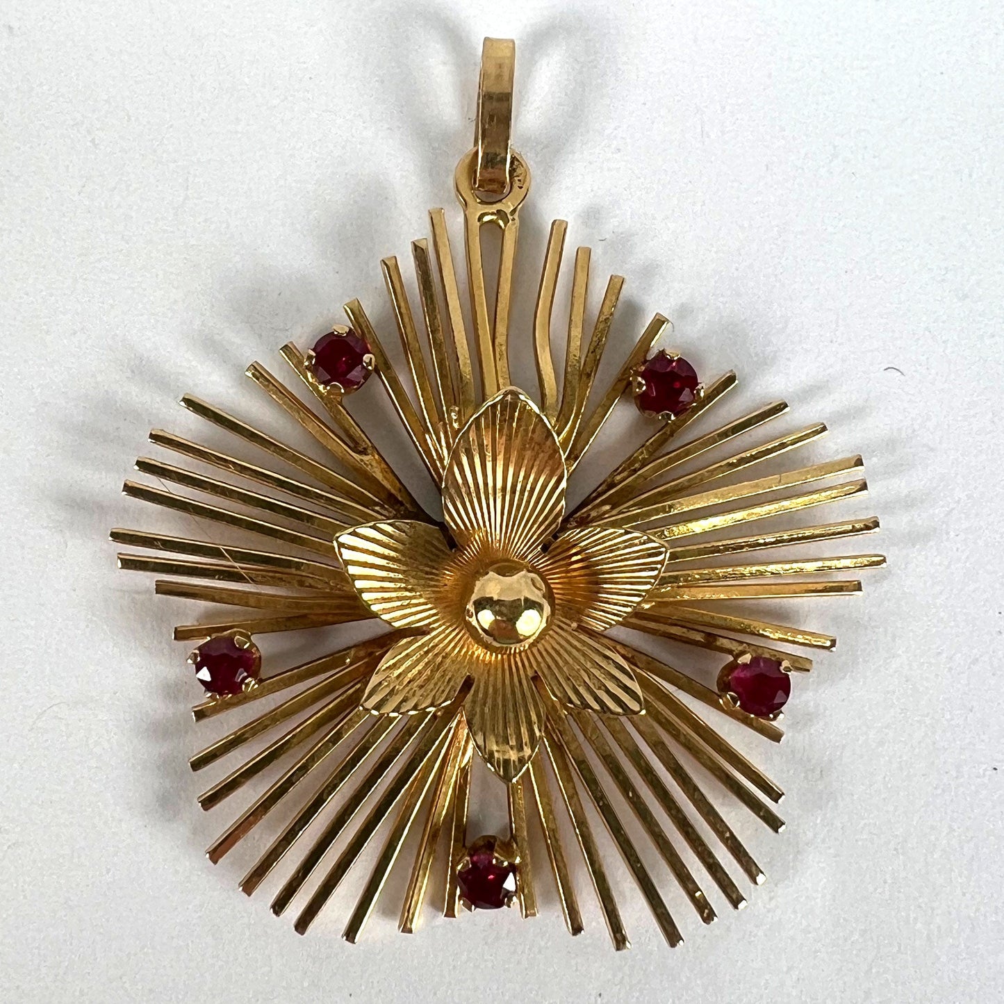 A yellow gold floral sunburst pendant with rubies, on a white background