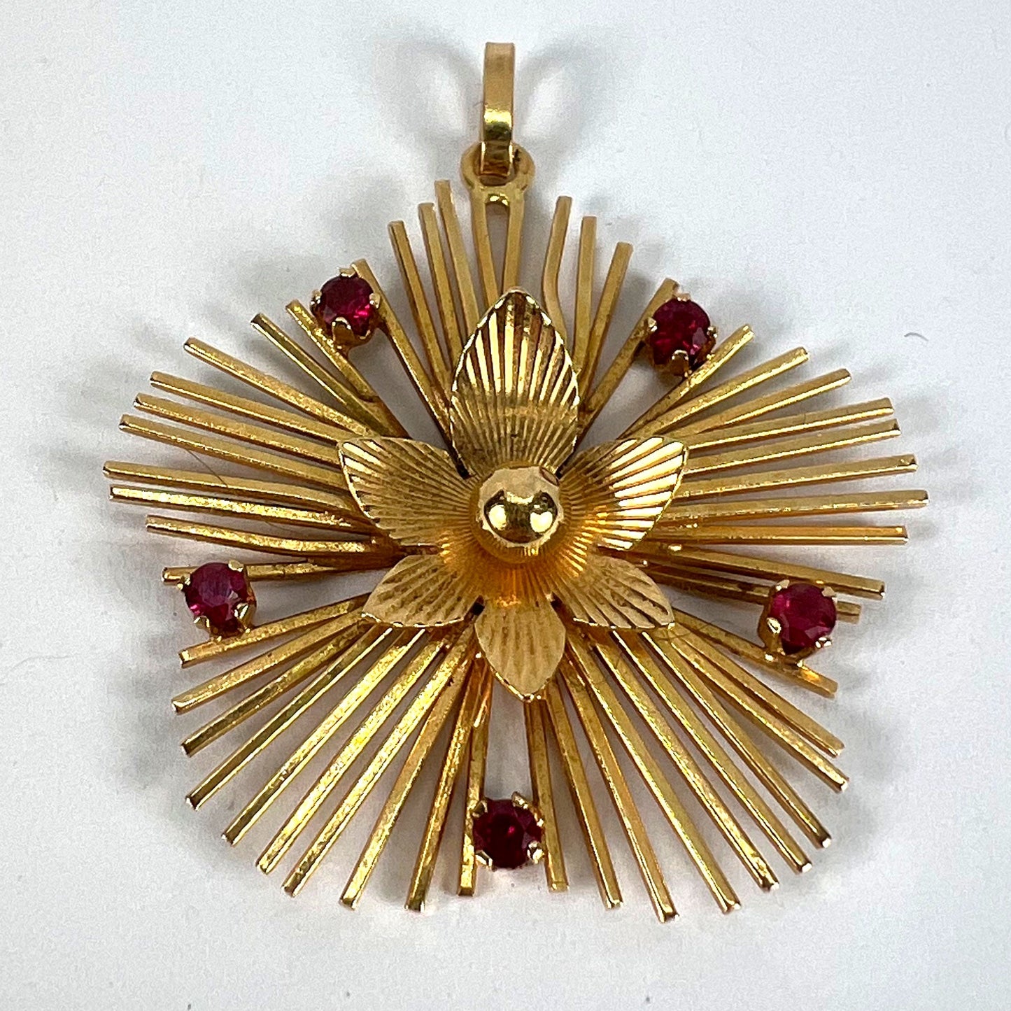 A yellow gold floral sunburst pendant with rubies, on a white background