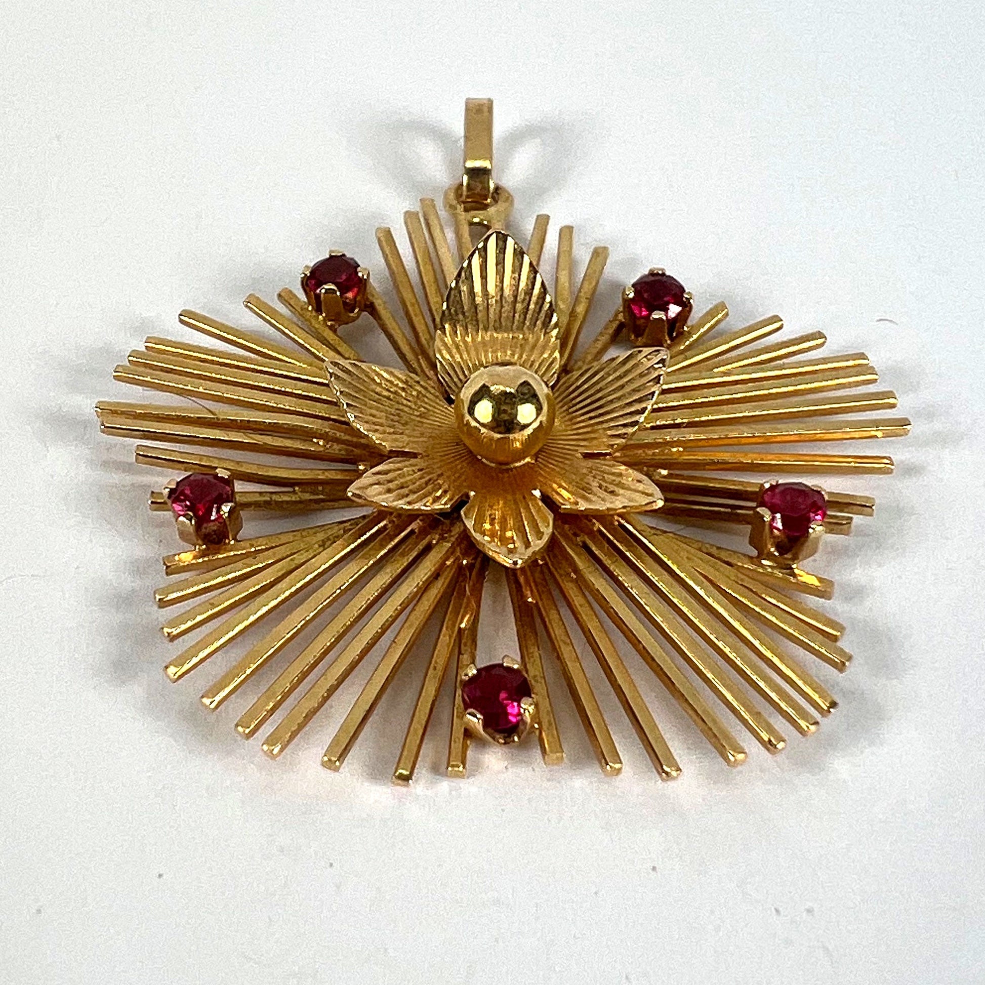A yellow gold floral sunburst pendant with rubies, on a white background