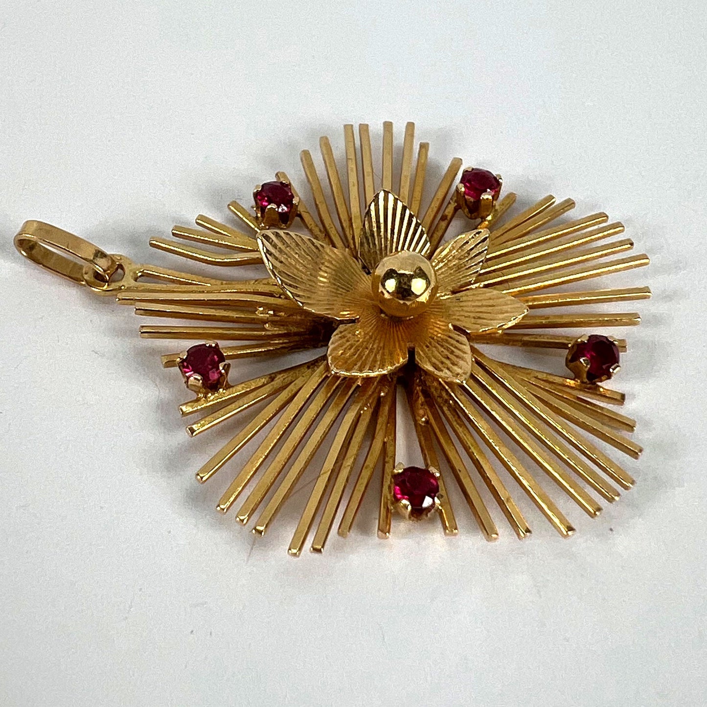 A yellow gold floral sunburst pendant with rubies, on a white background