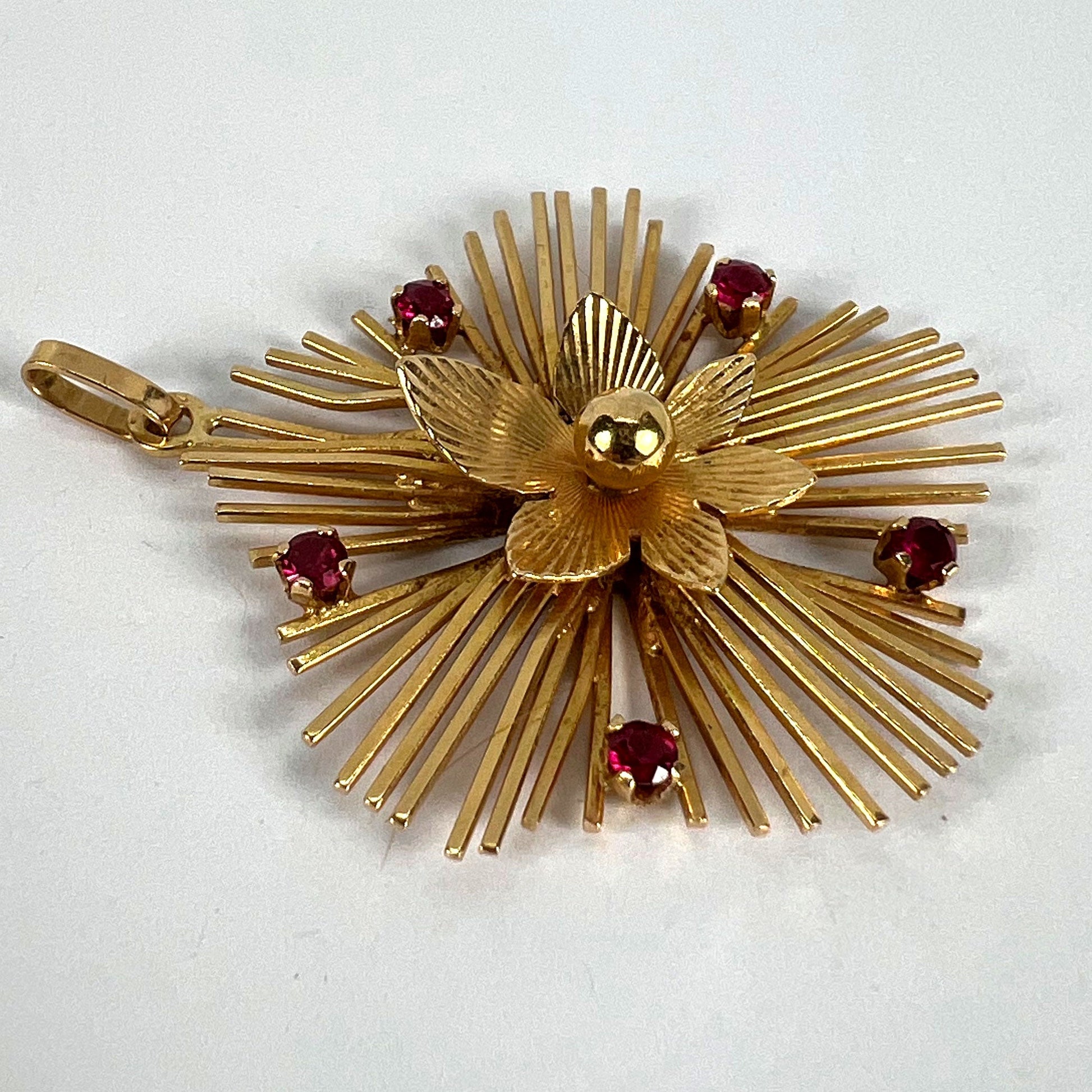 A yellow gold floral sunburst pendant with rubies, on a white background