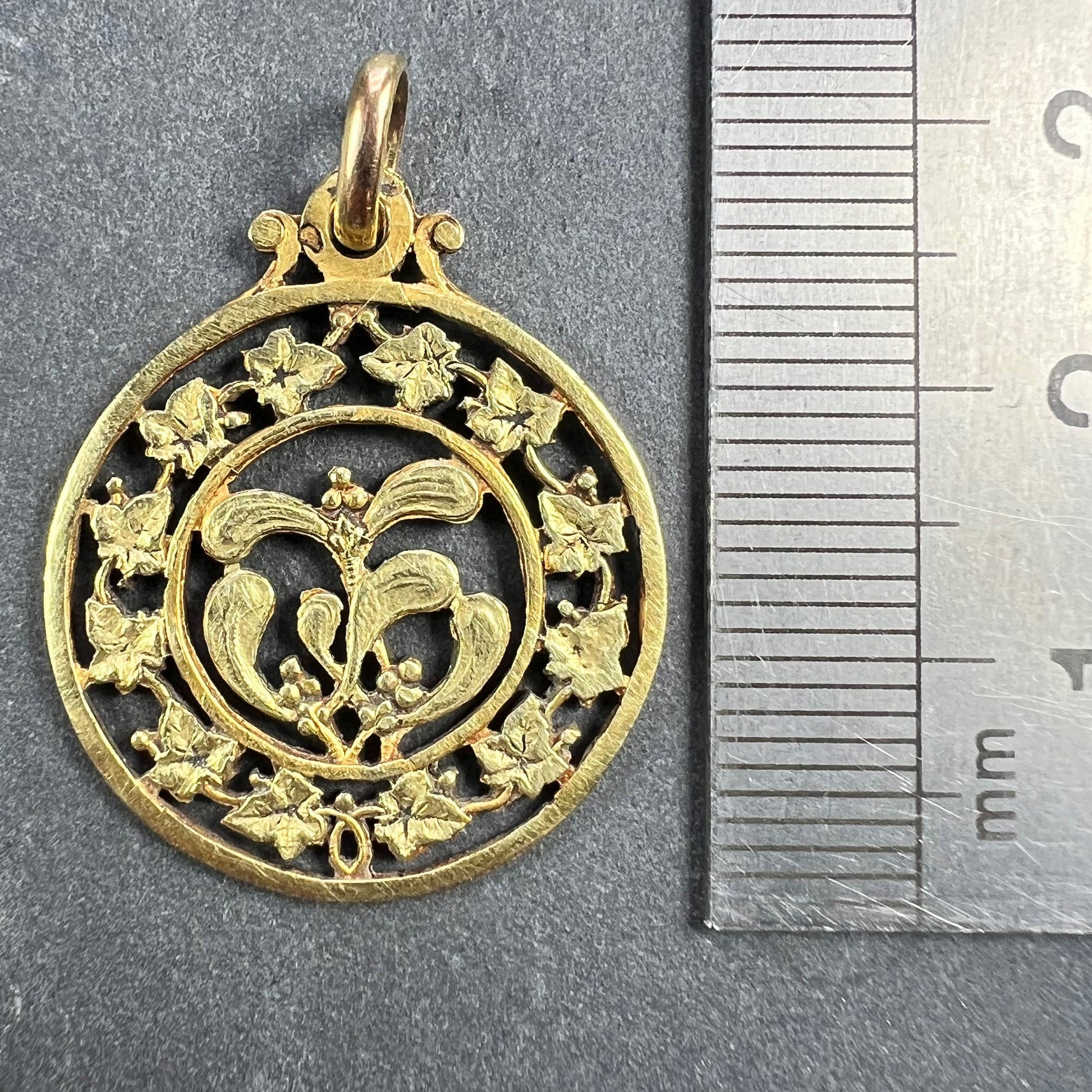 A round yellow gold pendant with mistletoe and ivy leaves, next to a metal ruler