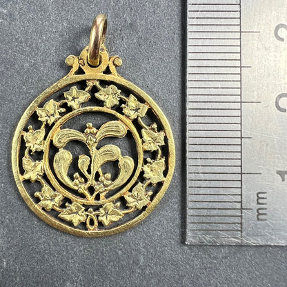 A round yellow gold pendant with mistletoe and ivy leaves, next to a metal ruler