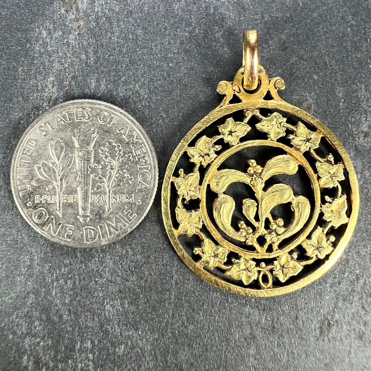 A round yellow gold pendant with mistletoe and ivy leaves, next to a dime coin