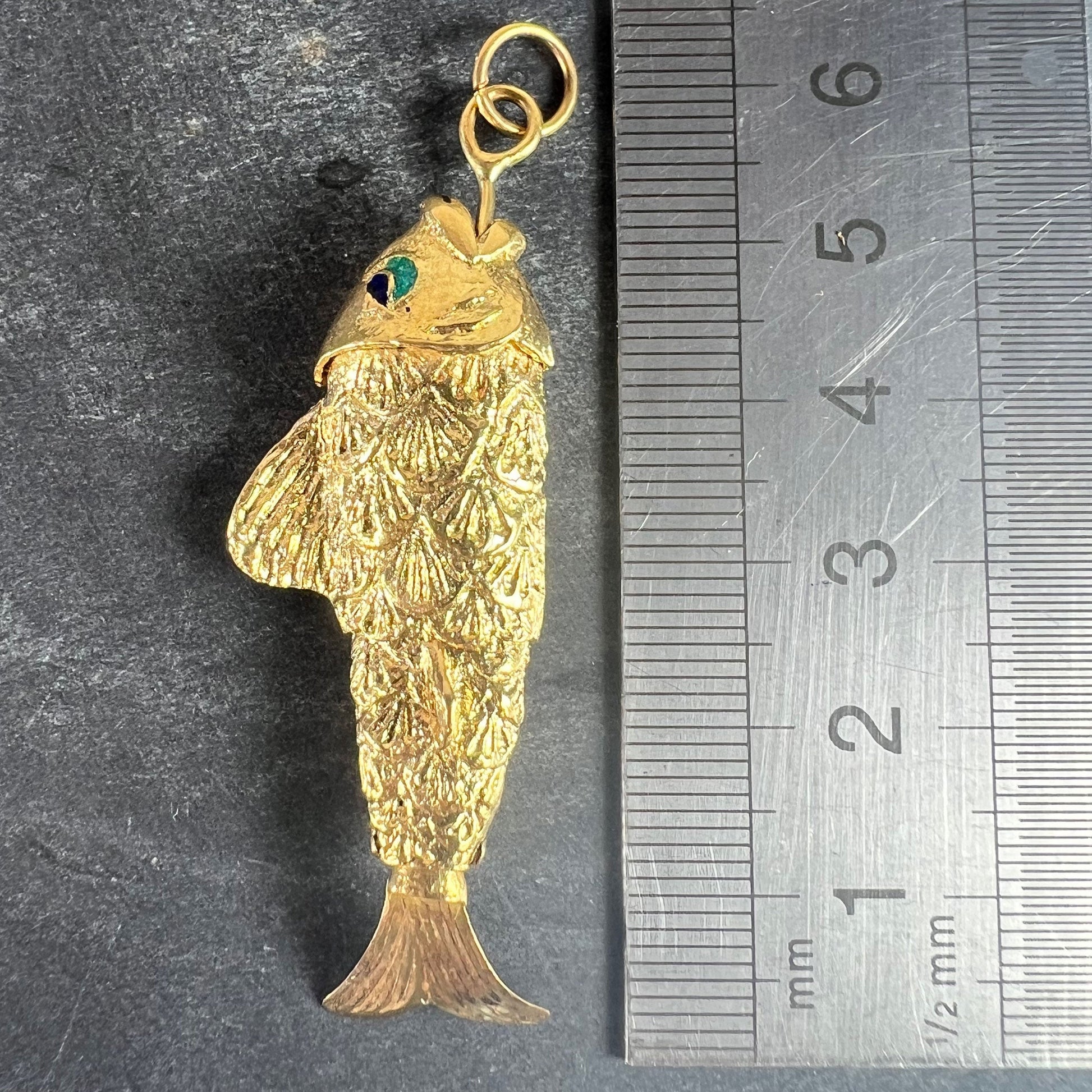 A yellow gold fish pendant with a green eye, next to a metal ruler